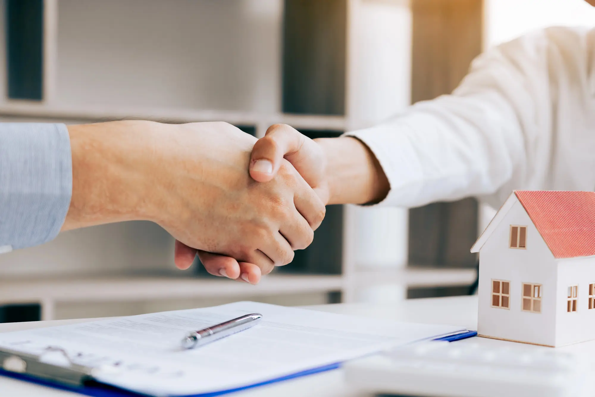 Two people shaking hands over a rental agreement with a miniature house model on the table, symbolizing a successful rent negotiation