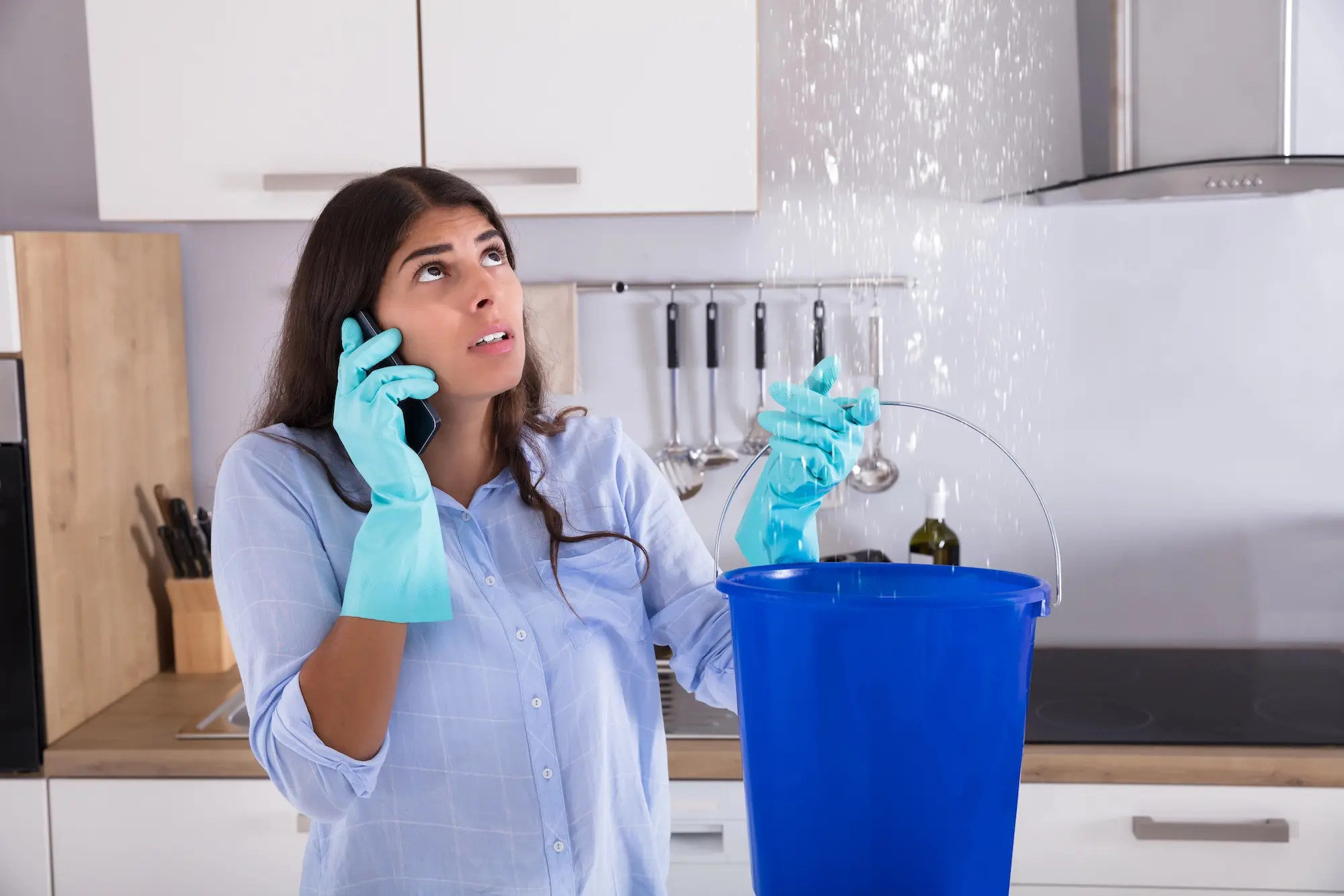 Tenant wearing gloves calls landlord while catching ceiling leak with bucket in kitchen