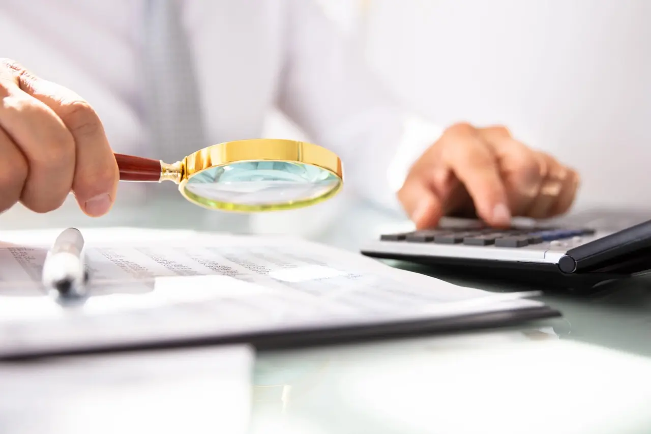 Person inspecting financial documents with a magnifying glass while calculating numberssymbolizing rental income verification and fraud detection
